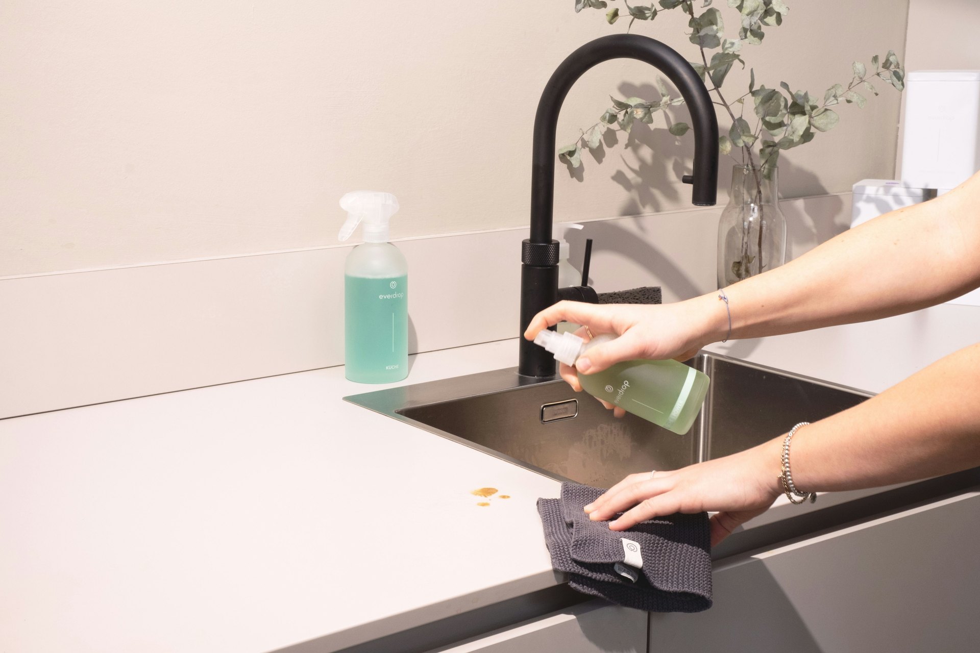 A person spraying cleaner and wiping a kitchen counter beside a black tap and sink