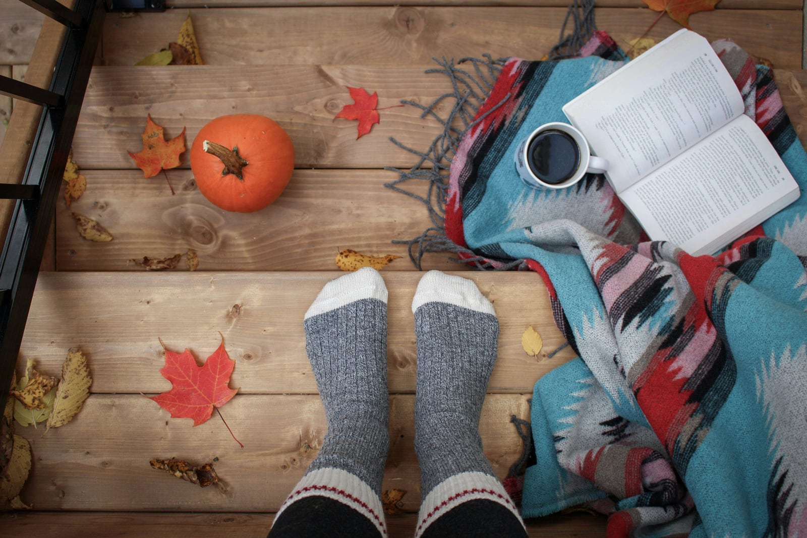 Wool-socked feet beside a pumpkin, autumn leaves, a blanket, an open book and a cup on wooden steps