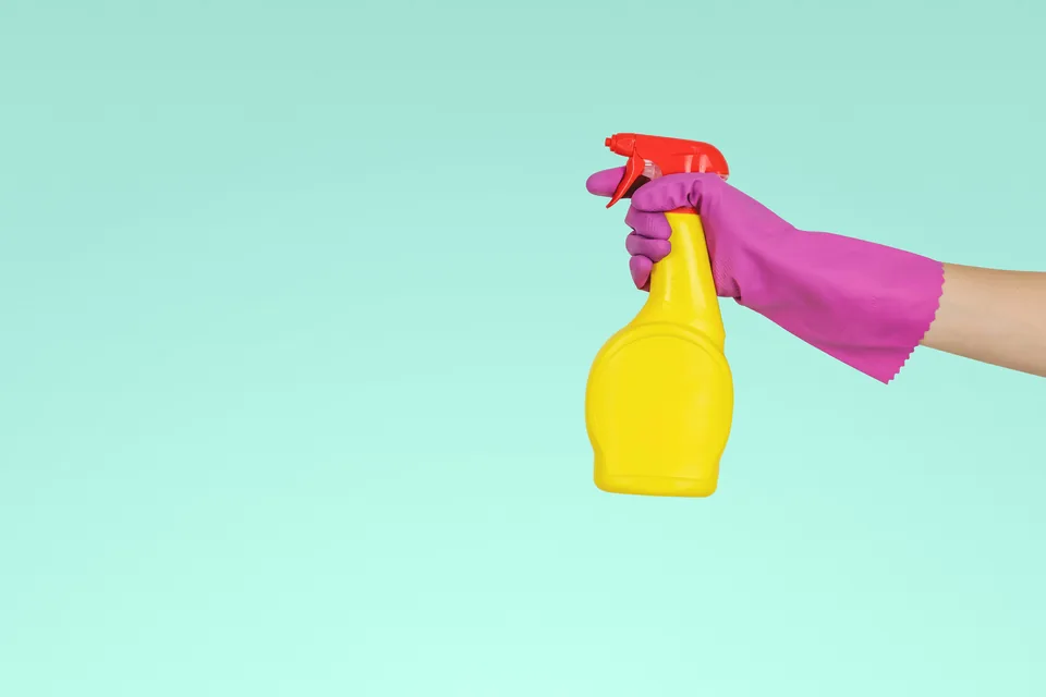 A hand in a purple cleaning glove holding a yellow spray bottle against a turquoise background